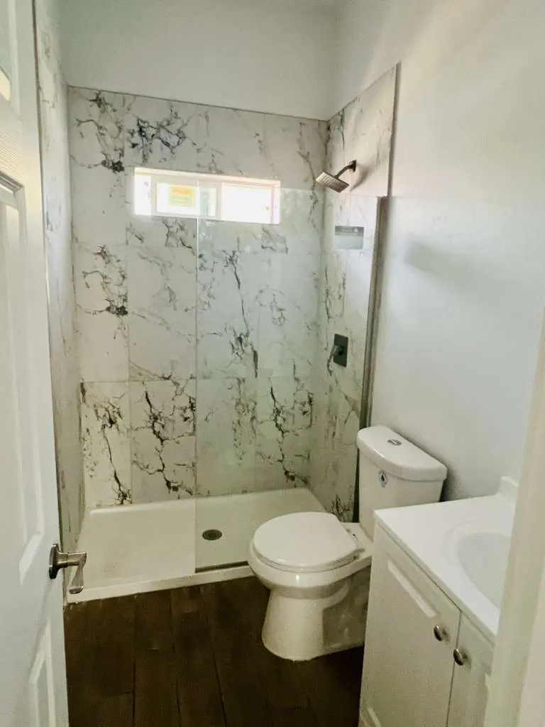 Baño completo con ducha de mármol, vanidad blanca y piso tipo madera oscura
