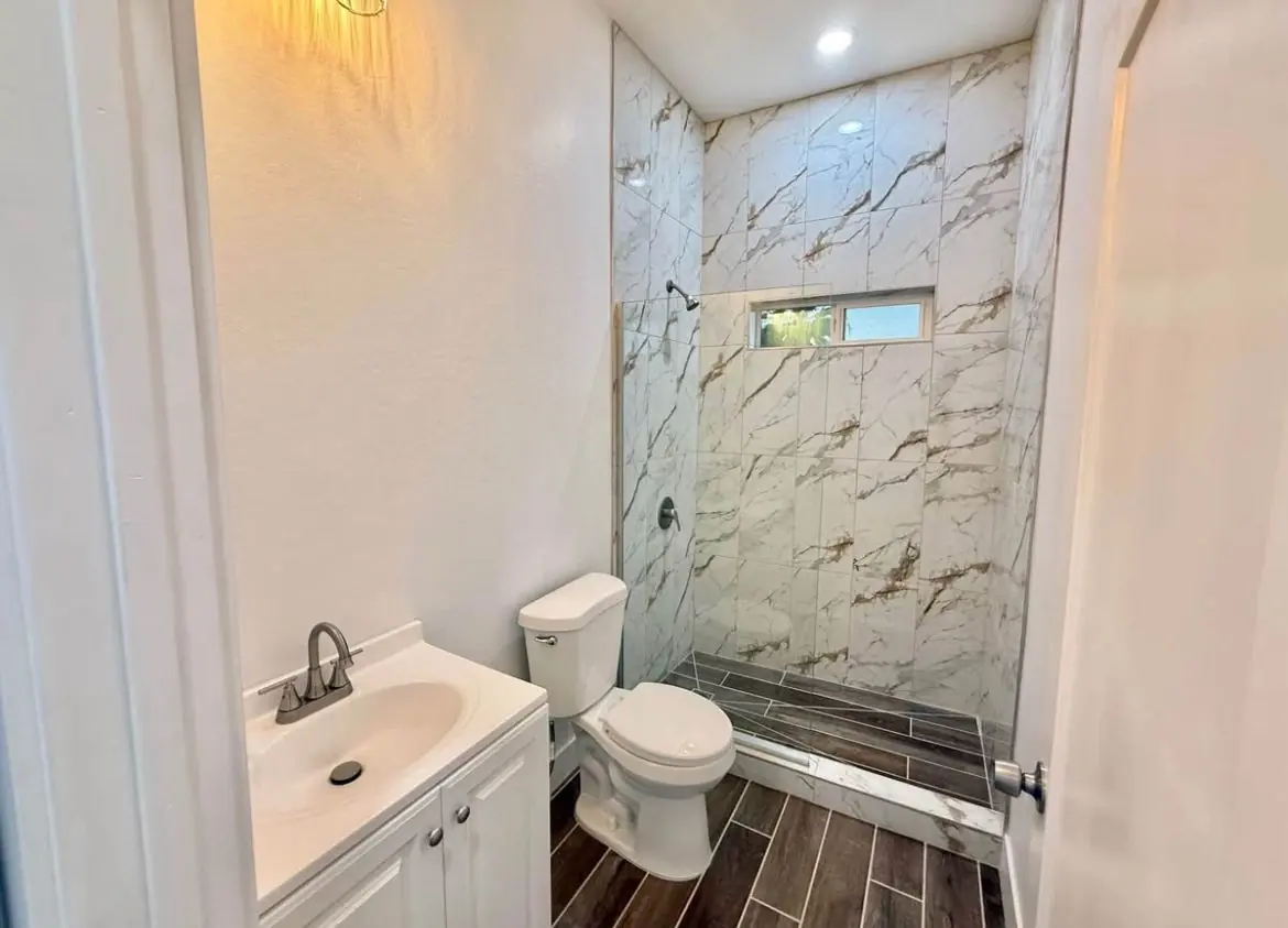 Baño completo con ducha de mármol, vanidad blanca y piso tipo madera oscura
